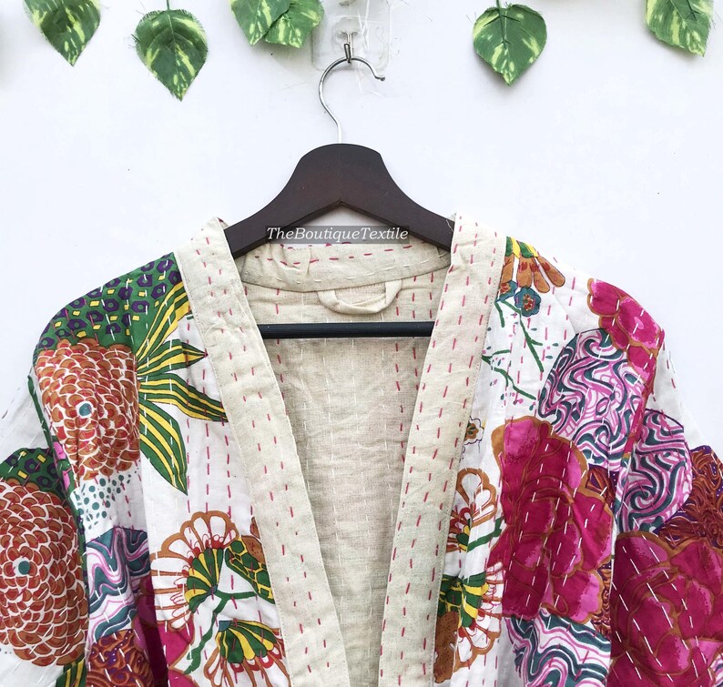 May include: A quilted jacket featuring a white base with vibrant floral and abstract patterns in pink, orange, green, and yellow. The garment has a beige collar and front trim with visible red running stitch details and the text TheBoutiqueTextile.