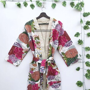 May include: A quilted cotton robe featuring a vibrant floral pattern with shades of pink, orange, and green on a white background. The garment includes long sleeves, a tie belt, two front pockets, and a cream-colored interior lining.
