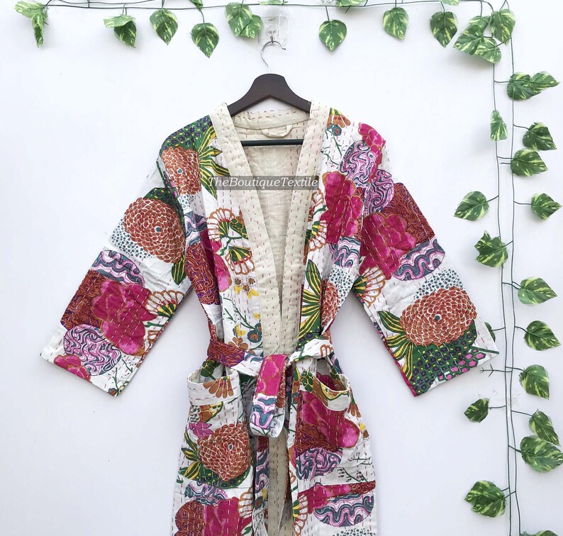 May include: A quilted cotton robe featuring a vibrant floral pattern with shades of pink, orange, and green on a white background. The garment includes long sleeves, a tie belt, two front pockets, and a cream-colored interior lining.