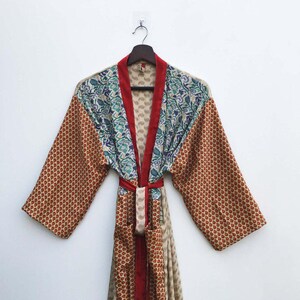 May include: A colorful, patterned robe with a red trim and belt. The robe features a mix of floral and geometric patterns in shades of red, blue, and beige. The robe is hanging on a wooden hanger against a white background.