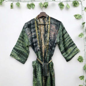 Könnte beinhalten: Ein Robe mit Batikmuster in Grün-, Schwarz- und Goldtönen hängt an einem Holzbügel. Die Robe hat einen Gürtel und ein gemustertes Innenfutter. Die Ärmel sind weit und der Stoff wirkt leicht.