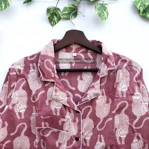 May include: A pink button-up shirt with a white print of stylized tigers. The shirt has a collar and long sleeves. The text "TheBoutiqueTextile" is printed on the shirt.