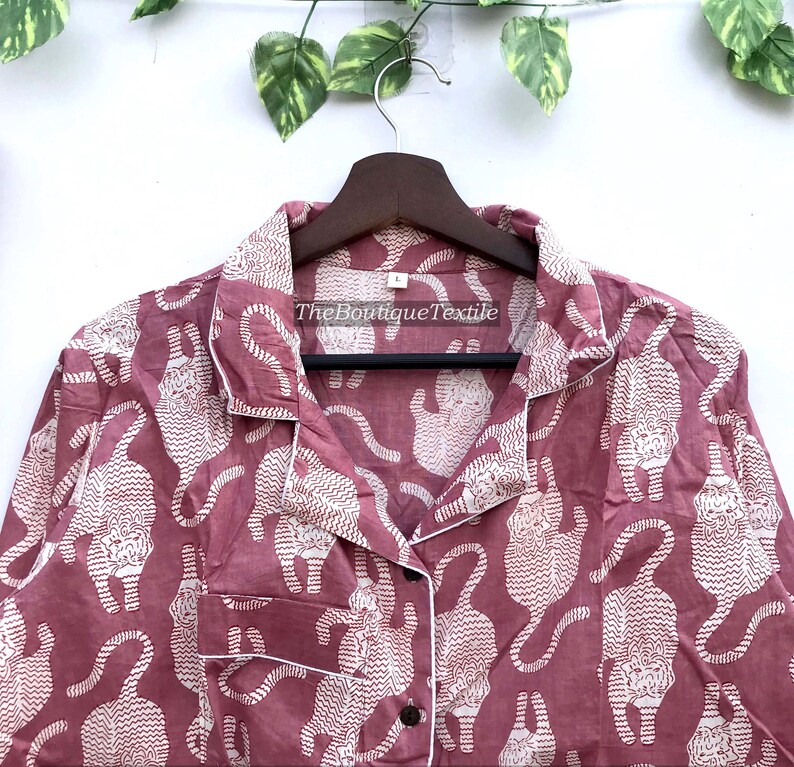 May include: A pink button-up shirt with a white print of stylized tigers. The shirt has a collar and long sleeves. The text "TheBoutiqueTextile" is printed on the shirt.