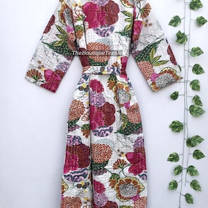 May include: A long-sleeved robe featuring a white base with a vibrant floral pattern in shades of pink, orange, green, and purple. The garment includes a matching tie belt, a cream-colored collar, and visible stitching throughout. Text reads TheBoutiqueTextile.