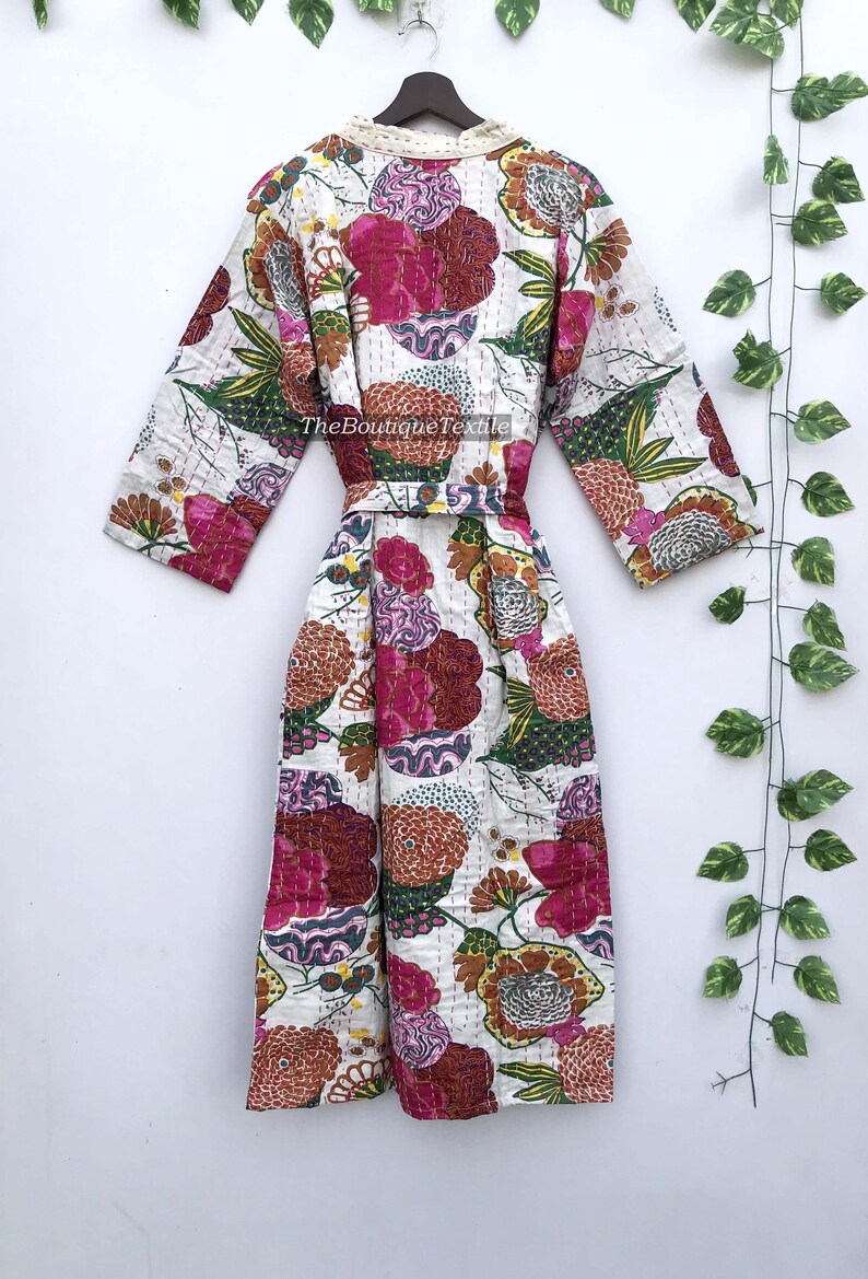 May include: A long-sleeved robe featuring a white base with a vibrant floral pattern in shades of pink, orange, green, and purple. The garment includes a matching tie belt, a cream-colored collar, and visible stitching throughout. Text reads TheBoutiqueTextile.
