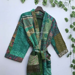 May include: A green patchwork robe with a tie belt. The robe is made of a variety of fabrics and has a quilted design. The robe is hanging on a hanger against a white wall.