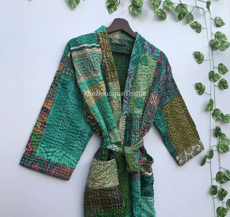 May include: A green patchwork robe with a tie belt. The robe is made of a variety of fabrics and has a quilted design. The robe is hanging on a hanger against a white wall.