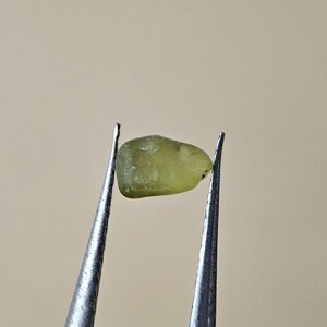 Peut inclure: Une petite pierre précieuse vert clair tenue par une pince argentée. La pierre précieuse a une forme arrondie et une surface légèrement texturée. La pince est positionnée sur un fond neutre, mettant en valeur la couleur et la forme de la gemme.