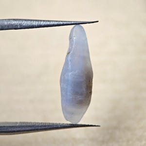 Puede incluir: Una gema alargada de color azul pálido es sostenida por unas pinzas. La gema tiene una superficie lisa y pulida con sutiles variaciones de color y textura. La gema es de un azul claro y translúcido.