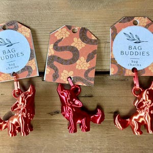 Puede incluir: Tres dijes para bolso con forma de lobo en metal rojo, sujetos a etiquetas de cartón rectangulares. Cada etiqueta tiene un círculo con el texto BAG BUDDIES y una ilustración botánica. Las etiquetas tienen un patrón de serpientes y flores.