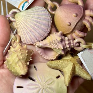 May include: A collection of sea-themed charms, including a crab, starfish, seashells, and a sand dollar. The charms are in shades of yellow, pink, and peach, with attached loops for hanging. The tag reads "BAG BUDD... charms".