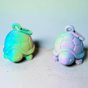 Könnte beinhalten: Zwei schildkrötenförmige Schlüsselanhänger mit Farbverlauf. Einer ist türkis und grün, der andere lavendel und blau. Jeder hat einen Ring zum Befestigen an Schlüsseln oder einer Tasche. Die Schlüsselanhänger liegen auf einer hellgrauen Oberfläche.