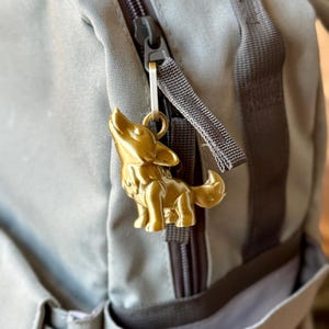 Może przedstawiać: Złoty wisiorek w kształcie wilka wisi na szarej zasuwce plecaka. Urok jest szczegółową, trójwymiarową figurą wyjącego wilka. Plecak jest w kolorze jasnoszarym z ciemnoszarymi akcentami.