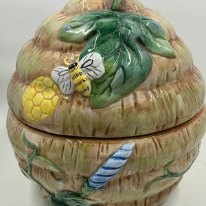 May include: A ceramic beehive-shaped container with a lid and handle. The container is painted with a brown, yellow, and green color scheme, featuring a bee, honeycomb, and leaves. The handle is a curved brown shape.