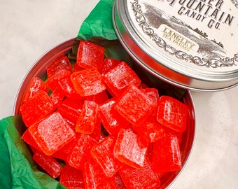 May include: A round metal tin filled with red, square-shaped hard candies. The tin is labeled "Linder Mountain Candy Co. Langley Tea Farms."
