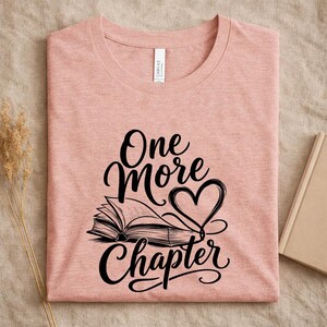 May include: A heathered peach-colored t-shirt featuring a black graphic print of an open book and a heart shape. The text "One More Chapter" is written in a stylized black script font above and below the book illustration on the front.