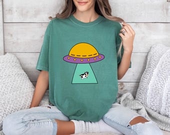 May include: A short-sleeve crewneck t-shirt in a muted green color. The center of the shirt features a graphic illustration of a yellow and purple UFO with a light green tractor beam shining down on a small black and white spotted cow.
