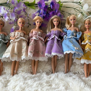 Peut inclure: Six poupées Barbie vêtues de robes faites à la main aux couleurs pastel. Chaque robe présente des broderies florales et des bordures en dentelle. Les poupées sont disposées sur une surface blanche et moelleuse, avec des fleurs violettes en arrière-plan.
