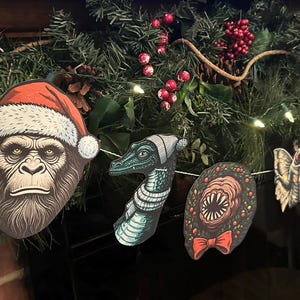 May include: A Christmas garland with five festive ornaments. The ornaments feature a creature from the black lagoon, a moth, a creature with a Santa hat, a creature with a scarf, and a creature with a bow tie.