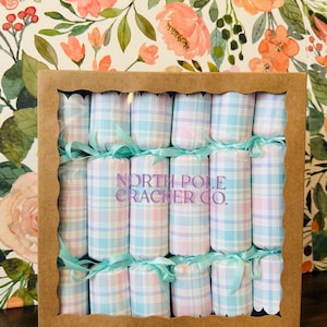 May include: A box of Christmas crackers with a plaid pattern in pastel blue, pink, and white. Each cracker is tied with a light blue ribbon. The box is brown with a scalloped edge. The text "NORTH POLE CRACKER CO." is printed on the crackers.