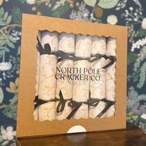 Op de afbeelding: Een doosje met kerstcrackers met een doorzichtig venster. De crackers zijn wit met gouden krullen en vastgebonden met zwarte linten. De doos is bruin en heeft de tekst "NORTH POLE CRACKER CO."