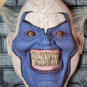 May include: A close-up of a creepy clown mask with a wide, toothy grin. The mask is painted blue and white with exaggerated features. The clown has orange eyes and a white, wrinkled forehead.
