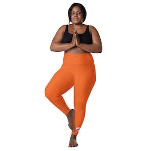 May include: A woman wearing a black sports bra and orange yoga pants with a small logo on the left leg. She is standing on one leg with her arms crossed in front of her chest.