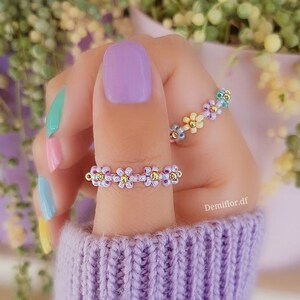 Può includere: Due anelli di perline con motivi floreali. L'anello sull'indice è fatto con perline rosa pastello, gialle e blu. L'anello sul pollice è fatto con perline viola e argento.
