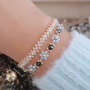 May include: Two beaded bracelets are shown, one above the other. The top bracelet is gold with iridescent beads. The bottom bracelet has gold beads with light blue flower-shaped beads and black beads. The bracelets are on a wrist, with a light blue sweater in the background.