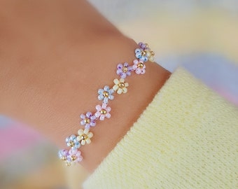 May include: A delicate bracelet made of small, colorful beads in shades of pink, yellow, and blue, arranged in a flower pattern. The bracelet is on a person's wrist.