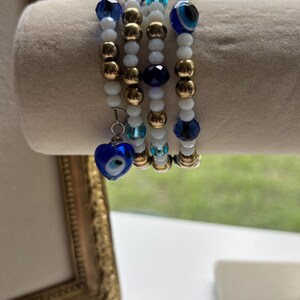 Könnte beinhalten: Ein mehrsträngiges Armband mit weißen, goldenen und blauen Perlen. Das Design umfasst facettierte dunkelblaue Perlen, metallische goldene Kugeln und kleine weiße runde Perlen. Ein herzförmiger blau-weißer Anhänger hängt am Armband.