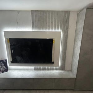 May include: A modern entertainment center with a flat screen TV mounted on a white wall. The wall is decorated with a marble-like pattern and vertical wood panels. The entertainment center has a gray countertop and two cabinets.