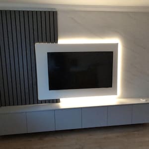 May include: A modern entertainment center with a white framed television mounted on a wall with gray wood paneling and white marble. The television has a white backlight. The entertainment center has a gray cabinet with three drawers.