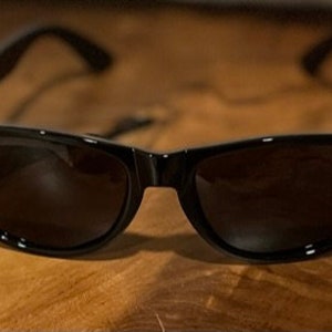 May include: Black plastic sunglasses with dark lenses. The sunglasses are on a wooden surface.