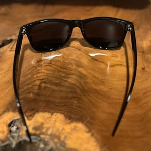 May include: Black sunglasses with a classic rectangular frame and dark lenses. The sunglasses are lying on a wooden surface.
