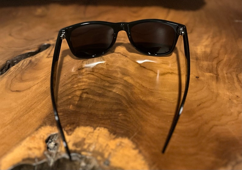May include: Black sunglasses with a classic rectangular frame and dark lenses. The sunglasses are lying on a wooden surface.