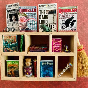 Puede incluir: Una estantería de madera en miniatura con nueve compartimentos, llena de libros en miniatura, una escoba, una botella de poción y otros objetos mágicos. Los libros incluyen títulos como "Harry Potter", "El Señor Oscuro" y "El Quisquilloso".