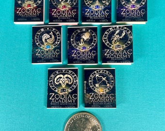 May include: Twelve miniature book covers with gold accents and black backgrounds. Each cover features a different zodiac sign with a title related to the sign. The titles include "The Awakening", "Rutebeis Fat", "The Rickgening", "Shadow Prince", "Curate", "Fatio Throne", "Heartless Sky", "Sorrow and Starlight", "Restless Stars", "The Awakening", "Rutebeis Fat", "The Rickgening".