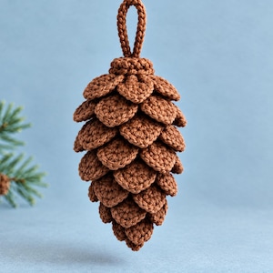Op de afbeelding: Een gehaakte dennenappelornament in bruin met een gestructureerd, gelaagd schubbenontwerp en een klein lusje aan de bovenkant om op te hangen. Het object is gemaakt van garen met een consistent steekpatroon dat een dennenappel nabootst.
