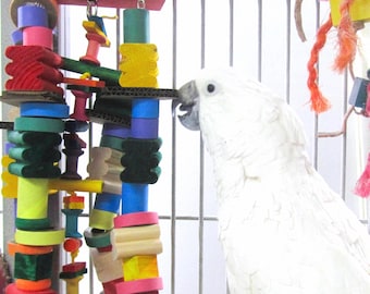 May include: A white cockatoo bird looking at a colorful wooden bird toy hanging in a cage. The toy is made of different shapes and sizes of wood, and is painted in bright colors. The bird is perched on a branch in the cage.