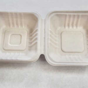 May include: A white, disposable, cardboard food container with a square shape and a slightly raised centre. The container is made of moulded pulp and is designed for single use.