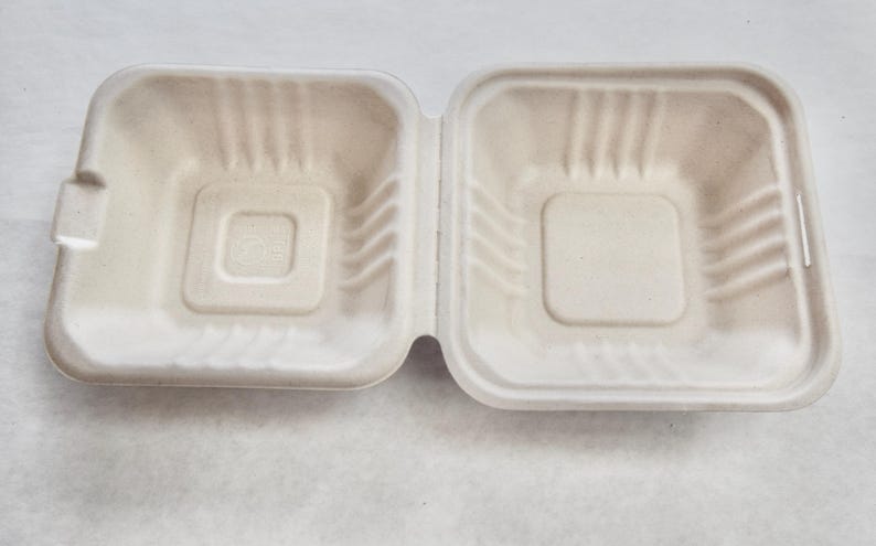May include: A white, disposable, cardboard food container with a square shape and a slightly raised centre. The container is made of moulded pulp and is designed for single use.
