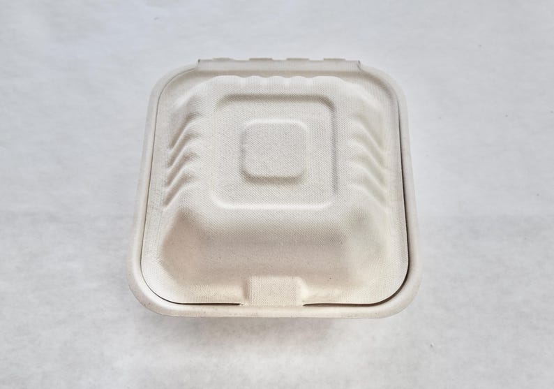 May include: A white cardboard food container with a hinged lid. The container is square shaped with rounded corners and a slightly raised centre.