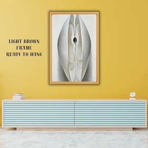 Puede incluir: Impresión de arte abstracto en un marco marrón claro, lista para colgar. La obra presenta una forma central blanca, similar a un pétalo, con un centro oscuro. El fondo es de un color suave y neutro. El texto "LIGHT BROWN FRAME READY TO HANG" es visible.