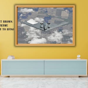 May include: A framed print showing an aerial view of a stealth bomber aircraft above clouds. The artwork is in a light brown frame and is ready to hang. The text on the frame says "LIGHT BROWN FRAME READY TO HANG".