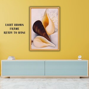 Pode incluir: Uma obra de arte emoldurada com três conchas em tons de castanho, bege e creme sobre um fundo branco. A obra de arte está numa moldura castanha clara e pronta para pendurar. O texto na parede diz "LIGHT BROWN FRAME READY TO HANG".