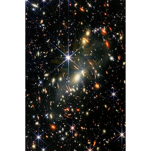 May include: A deep space photograph showing a dense field of distant galaxies and bright stars against a black background. The image features numerous points of light in shades of white, orange, and red, with some stars displaying distinct diffraction spikes.