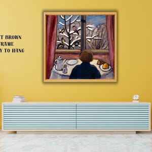 May include: A framed painting depicts a person gazing out a window at a snowy landscape with birds. The scene includes a table set with a teapot, plate, and food. The frame is light brown, and the text "LIGHT BROWN FRAME READY TO HANG" is on the wall.
