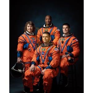 May include: Group portrait of four astronauts in orange and blue space suits. The suits feature NASA logos and patches. One astronaut holds a helmet. The background is a dark blue.