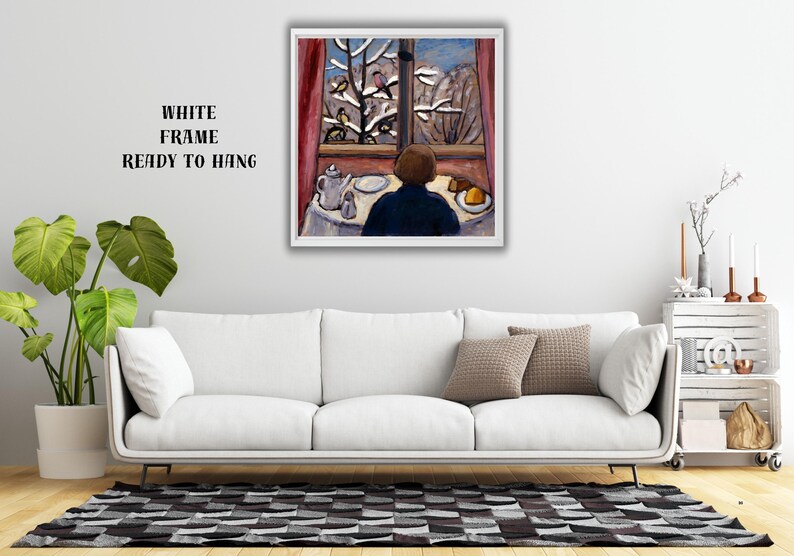 May include: A framed painting depicts a person seated at a table, gazing out a window at a snowy landscape with birds. The artwork features a white frame and the text "WHITE FRAME READY TO HANG". The painting is displayed above a white sofa.